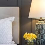 A styled nightstand with a patterned lamp and yellow flowers beside an upholstered bed