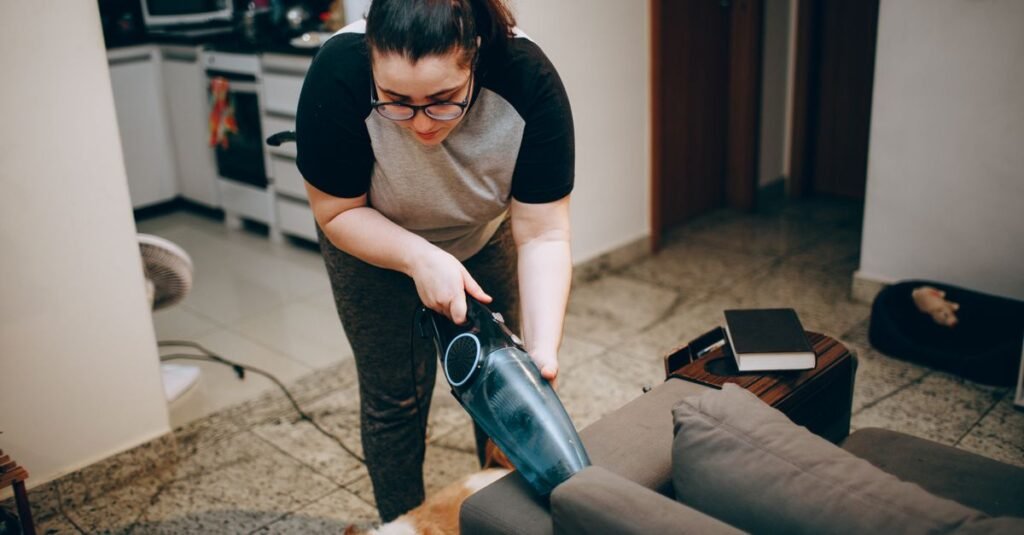 Woman vacuuming a sofa with a handheld unit while her dog watches