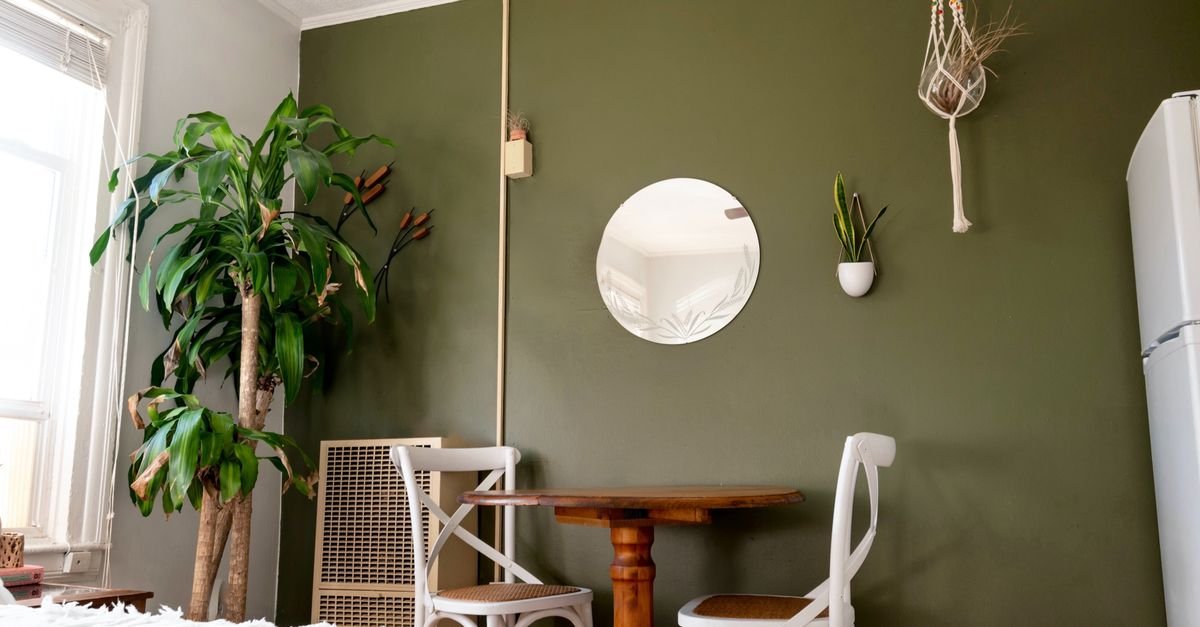 A round mirror on a green wall beside plants and a chair